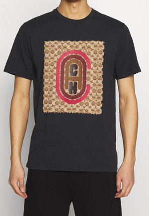 Man wearing black t-shirt with a large beige and brown patterned square featuring red and brown stylized "CAH" letters on the front.