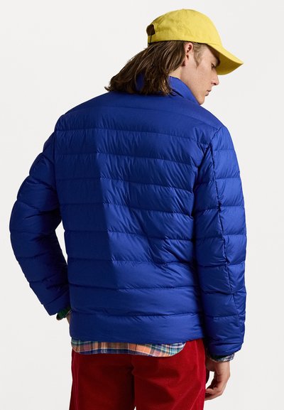 Blå pufferjakke med vandret quiltet design, med højt krave og vist fra ryggen. Båret over en mønstret skjorte.