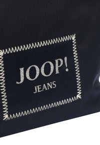 Námořnická textilní taška s stříbrným vyšitým nápisem "JOOP! JEANS," orámovaným bílým švem. Hladký povrch, minimalistický design.