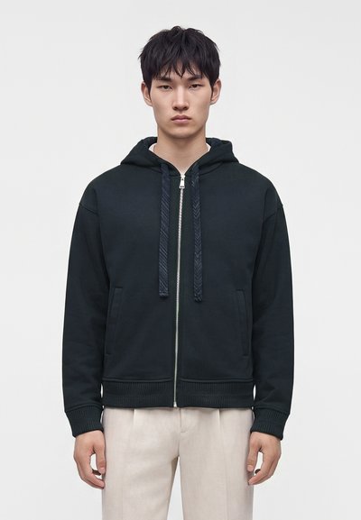 Jeune homme portant un sweat à capuche noir zippé avec cordons et pantalon beige, debout sur un fond clair uni.
