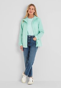 Light mint green windbreaker with a hood, zip closure, elastic cuffs, worn over a light blue shirt. Paired with high-waisted jeans and white sneakers.