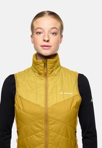 Isolierte ärmellose Jacke in senfgelb mit Steppmuster, Frontreißverschluss und Markenlogo. Schwarzes Langarmshirt darunter.