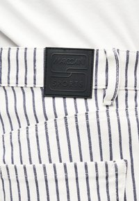 Λευκό ύφασμα με κάθετες ναυτικές ρίγες, που δείχνει μια πίσω τσέπη και μια μαύρη ετικέτα της μάρκας Marc Cain Sports πάνω από την τσέπη.