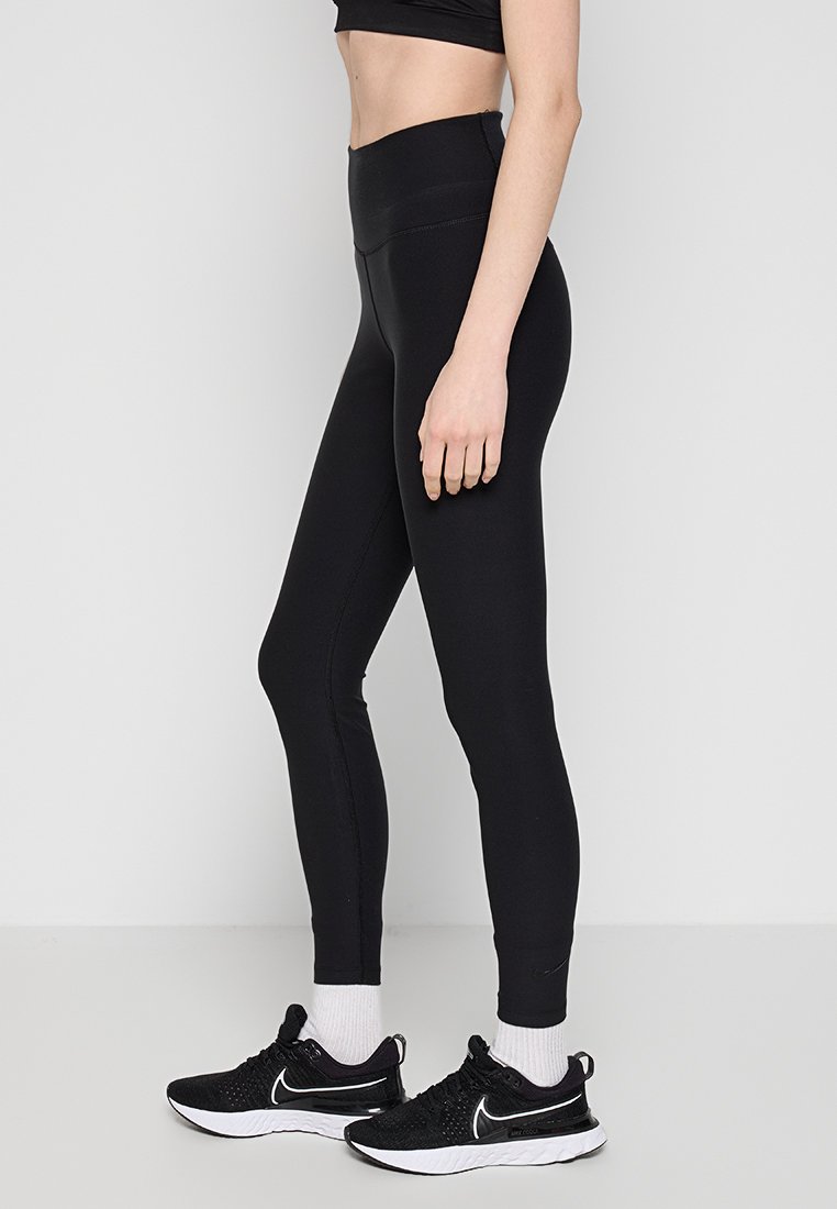 Leggings neri a vita alta realizzati in materiale elasticizzato, con una superficie liscia e un design aderente. Indossati con scarpe da ginnastica nere.