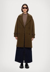Olive green double-breasted coat with wide lapels, paired with a fitted brown knitted sweater and dark denim maxi skirt. Black chunky shoes.