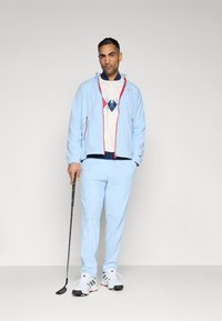 Chaqueta deportiva azul claro con detalles rojos, polo blanco con un patrón de diamantes, pantalones a juego y un palo de golf negro con zapatos blancos.