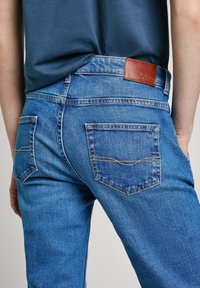 Blå denimjeans med medelvattning, som har två bakfickor och en brun läderlapp vid midjan, synliga sömmar.