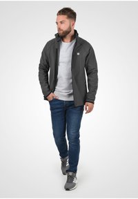 Heren grijze softshell jack met een hoge kraag, ritssluiting aan de voorkant en zakdetails, gecombineerd met een grijze sweatshirt en een blauwe spijkerbroek.