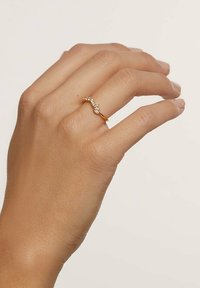 Hånd med guld ring, der har et buet design og fem små klare ædelstene, sat mod en enkel lys baggrund.