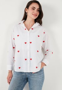 White button-up shirt made of lightweight fabric, featuring scattered red heart embroidery, long sleeves, and a classic collar design.