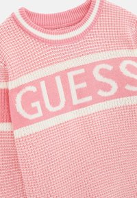 Maglione rosa a maglia con colletto rotondo, dettagli a strisce bianche e la parola "GUESS" ben visibile al centro.