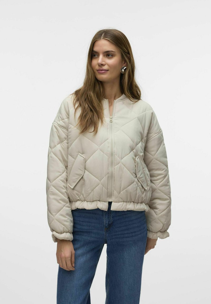 Kergelt beež padded bomber-jakk, millel on teemantkudumine muster, ribiga alläär ja lukuga sulgemine. Omab kahte küljetaskut ja ümarat kaela.