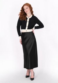 Cardigan nero cropped con accenti e bottoni crema, abbinato a una gonna midi nera in satin e scarpe con tacco alto nere.