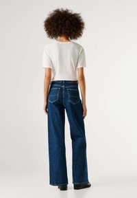 Široke modre kavbojke z nezačiščeno robom in poudarjenim šivom, kombinirane z belo oprijeto majico, videne od zadaj.