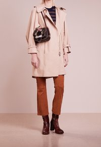 Femme blonde portant un trench-coat beige, un pull à rayures, un pantalon couleur rouille, des bottes texturées brun foncé, avec un sac à bandoulière noir décoré.