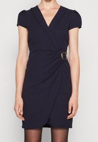 Femme portant une robe portefeuille bleu marine à manches courtes avec un décolleté en V profond et une boucle dorée à la taille, assortie à des collants noirs transparents.