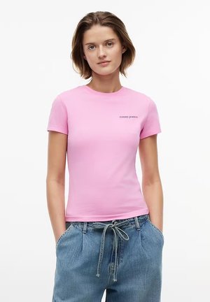 Donna con capelli castani corti che indossa una maglietta rosa aderente Tommy Jeans e jeans blu larghi con cintura annodata, mani nelle tasche.