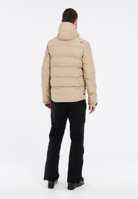 Doudoune beige avec capuche amovible, présentant des sections matelassées, portée sur un pantalon noir. Le design inclut des coutures visibles et des zippers.