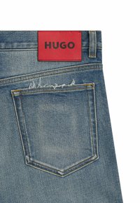 Farkut vaaleansinisinä, joissa on hieman kulunut takatasku, varustettuna punaisella nahkalaminoidulla tarralla, jossa on mustalla painettu "HUGO".