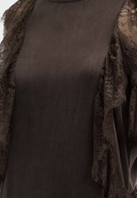 Σκούρο καφέ ύφασμα με διαφανείς βολάν από δαντέλα κατά μήκος των άκρων του ώμου και των μανικιών, που αναδεικνύουν μια υφή υφάσματος με ανάγλυφη λεπτομέρεια.