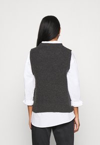 Chaleco de punto gris con textura ribeteada, que presenta un cuello alto y un diseño sin mangas, llevado sobre una camisa blanca de botones.
