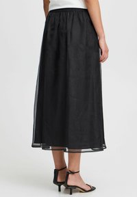 Gonna maxi nera e trasparente con vita elastica, caratterizzata da una texture morbida e una silhouette fluida. Indossata con tacchi a strappo neri.