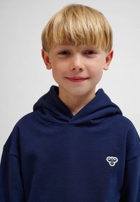 Marinblå luva med mjuk textur, som har ett litet broderat emblem på vänster bröstområde.