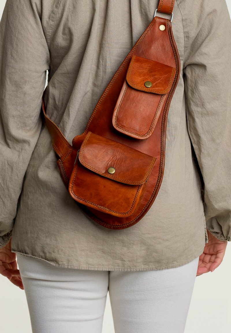 Brown leather sling bag with three front pockets, metal snaps, and a curved shape, worn over a light-colored shirt. Smooth texture.