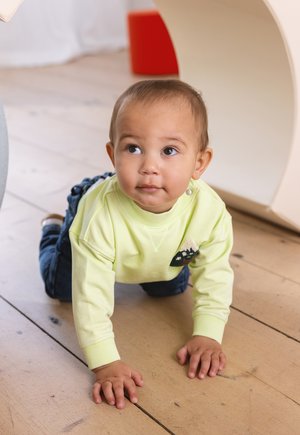 Kriechendes Kind in einem leuchtend gelben Sweatshirt mit einem Berg-Patch, kombiniert mit blauen Jeans, auf einem hellen Holzboden.