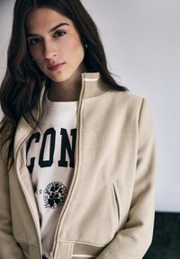 Veste zippée beige en tissu texturé, col montant et accents rayés. Portée par-dessus un t-shirt blanc avec des inscriptions noires et un détail graphique.