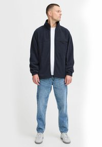 Marineblå fleecejakke med høy krage, frontlomme med glidelås og lange ermer. Brukt over en hvit t-skjorte og kombinert med lyseblå jeans.