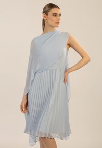 Robe plissée bleu clair avec un superposé drapé asymétrique. Fabriquée dans un tissu doux et fluide, elle présente un design sans manches et une longueur jusqu'aux genoux.