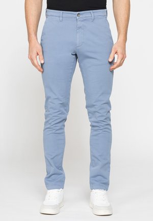 Uomo con pantaloni slim fit azzurri chiari, sneakers bianche e una maglietta nera, in piedi contro uno sfondo chiaro e semplice.
