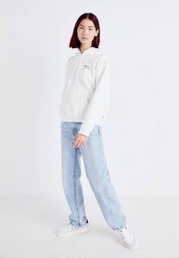 Vit hoodie med en ficka fram, dragsnöre på huvan och en liten färgglad logga. Kombinerad med ljusblå raka jeans och vita sneakers.