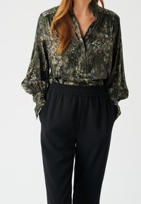 Blouse boutonnée vert foncé à imprimé léopard, avec des manches longues bouffantes. Associée à un pantalon noir à taille élastique. Texture de tissu lisse.