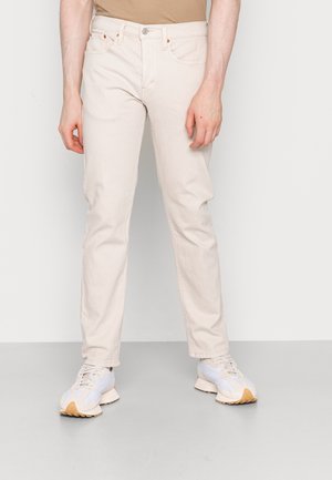 Personne portant un jean beige slim fit et des baskets blanches, debout devant un fond clair uni.