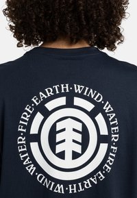 Camiseta de algodón azul marino con un gran gráfico circular en la parte posterior que lleva las palabras "AGUA", "FUEGO", "TIERRA" y "VIENTO" en blanco.