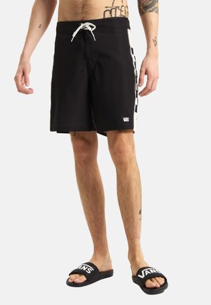 Uomo che indossa pantaloncini neri Vans con strisce laterali bianche e ciabatte Vans nere a piedi nudi su sfondo bianco.
