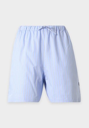Femme portant une chemise ample à rayures bleues boutonnée sur un tee-shirt blanc, avec un short rayé bleu assorti et des ballerines noires pointues, debout, mains sur les hanches.