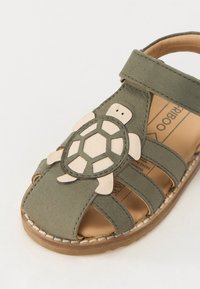 Grønn skinnsandale med et skilpadde-design. Har utskjæringer, en stropp, og en brun såle. Merkbar lys beige skilpadde-pynt.