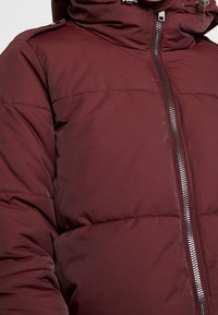 Giacca invernale bordeaux con collo alto, chiusura a zip e sezioni a quadri orizzontali, realizzata in tessuto liscio e leggero.