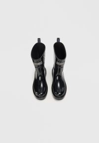 Bottes de pluie en caoutchouc noir avec une finition lisse et brillante. Hauteur mi-mollet, bout arrondi, doublure en textile et languettes de tirage. Motif crocodile sur les côtés.