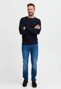 Mørkeblå strikket genser med rund hals, kombinert med lyseblå jeans og svarte sko. Genseren har et lite logo på brystet.