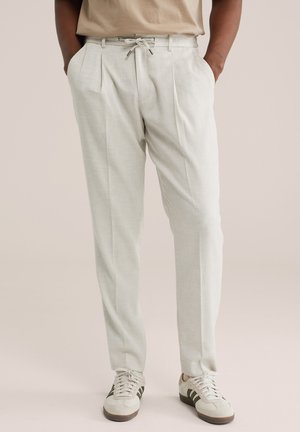 Homme portant un pantalon plissé beige clair avec taille à cordon et des baskets blanches à rayures foncées, les mains dans les poches.