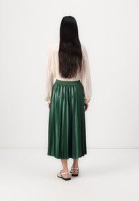 Zelená saténová sukně s plissé a vysokým pasem, spárovaná se světlou, strukturovanou blůzou s dlouhým rukávem. Obuv tvoří sandály na řemínky.