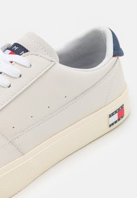 Zapatilla de cuero blanca con cordones planos blancos, detalles de costura y suela texturizada color crema. Presenta un acento de marca en azul, rojo y blanco.