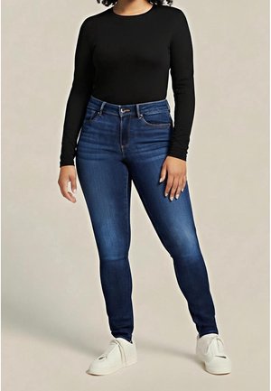 Frau trägt ein schwarzes langärmeliges Oberteil, dunkelblaue enge Jeans und weiße Slipper, steht vor einem einfarbigen Hintergrund.