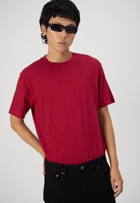 Camiseta de algodón burdeos con mangas cortas, escote redondo y un sutil detalle de logo. Combinada con jeans negros y gafas de sol.