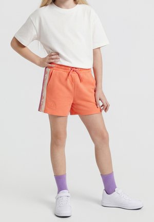 Dziecko ubrane w białą koszulkę, pomarańczowe spodenki z bocznymi paskami z logo, fioletowe skarpetki i białe trampki, stoi z jedną ręką na biodrze.