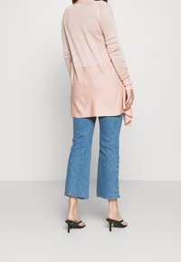 Ljusrosa långärmad cardigan med texturerad baksida, parat med högmidjade blå utsvängda jeans och svarta klacksandaler.